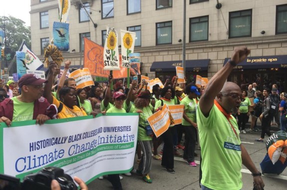 a group of black people coming together to march in a protest against climate change. their hands are raised in the air and they hold a banner that reads historically black colleges and universities climate change initiative