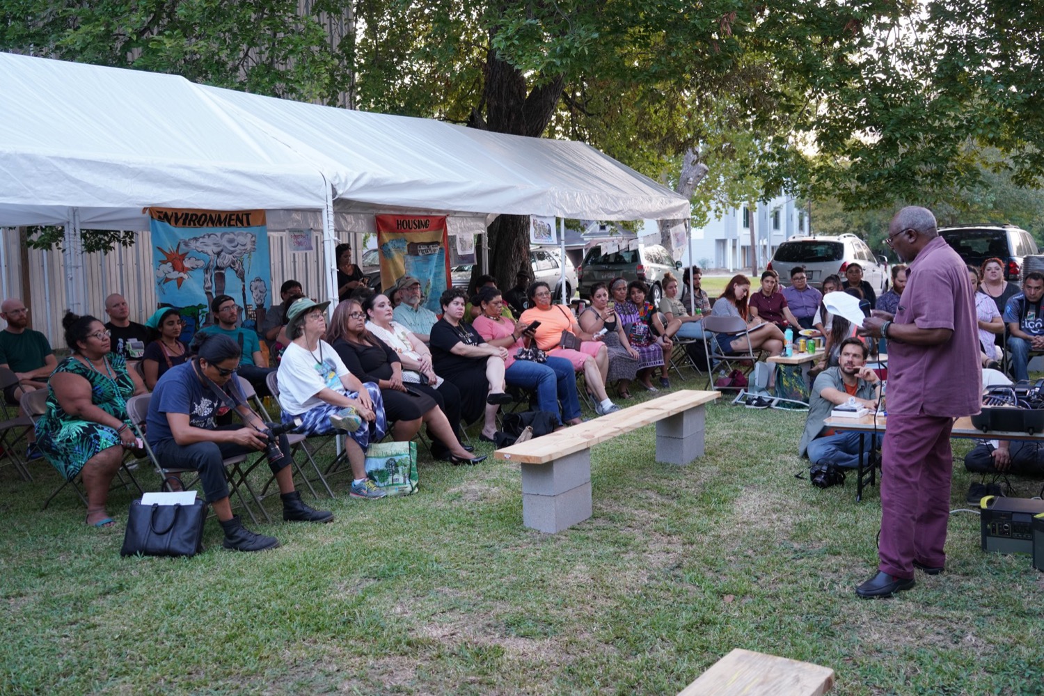 a black man wearing all purple speaks to a crowd of people under an outdoor tent