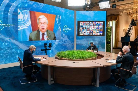 three men in face masks sit around a circular table and speak with people on a zoom call up on a large screen