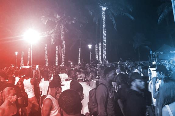 a large throng of people at night, tightly packed without masks. a street sign is in the foreground with palm trees decorated with string lights in the background