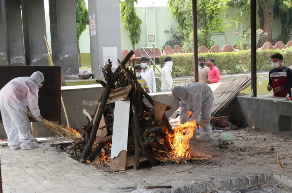 two people in hazmat suits attends to bonfire that is cremating someone who'd died from covid-19 complications in an abandoned building. people wearing masks observe from a distance