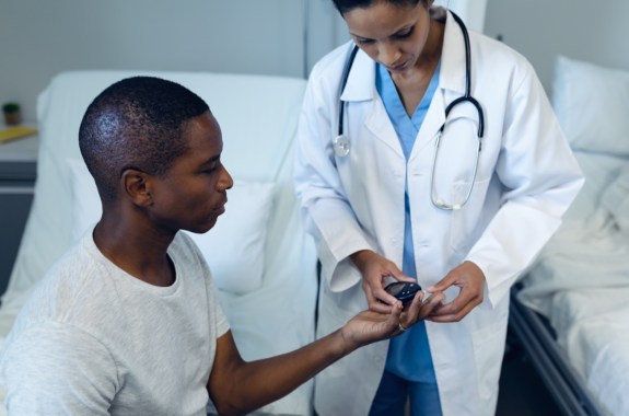 Side view of mixed race female doctor taking black patient's blood sample with lancet pen in the ward at hospital