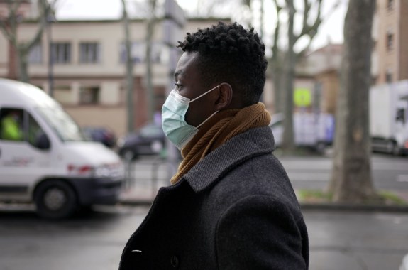 a black person wearing a mask on the street with cars and buildings behind them