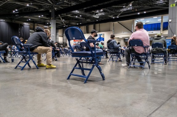 people sitting in rows of seats in large indoor stadium wearing masks. a chair in the foreground is empty