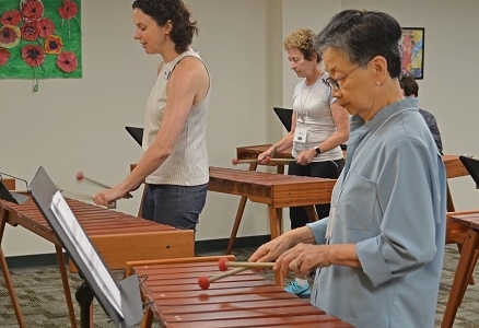 a group of people playing xylophones in a class, with the main focus on an older asian woman with short hair