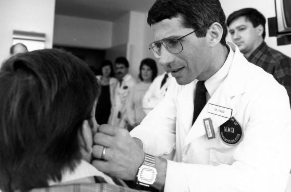 a black and white photo of a male doctor with glasses wearing a white coat with a patch that reads NIAID is examining a patient whose back is to the camera. other clinicians are in the office watching him treat the patient