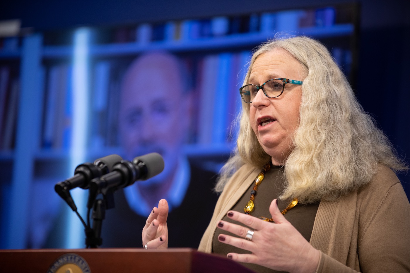 a white woman speaking at a podium with a microphone with a projected image of a white man behind her