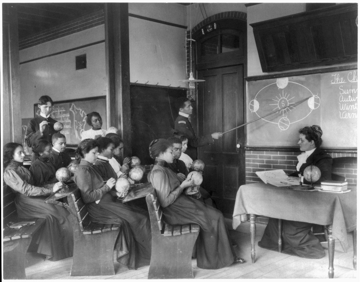 a very old black and white photo of a classroom with over a dozen adult students. most of the students are black, and each of them are holding a miniature globe of the earth. a black man, using a long pointing stick, is gesturing towards the blackboard, where a simple chalk diagram is drawn depicting the different phases and tilts of the earth as it orbits around the sun