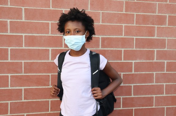 black kid wearing mask stands in front of brick wall with a backpack on