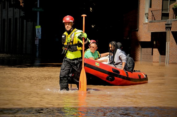 a white man in a hazard vest, hard hat, and water pants holds an oar with one hand and pulls a small inflatable rescue boat with four people in it through a flooded street. the water goes up to his shins