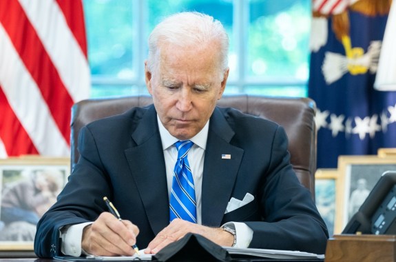 an older white man with white hair in a suit signs paperwork at a desk with the american flag behind him