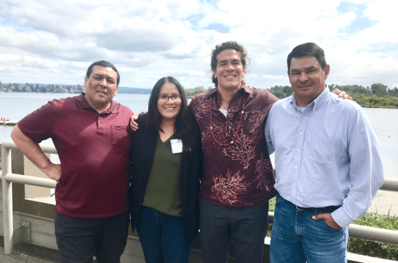 four native americans, three men, one woman, stand next to each other smiling at the camera, with a lake and lush landscape behind them
