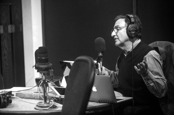 a black and white image of a man in glasses in a studio. he has headphones on and is speaking into a microphone