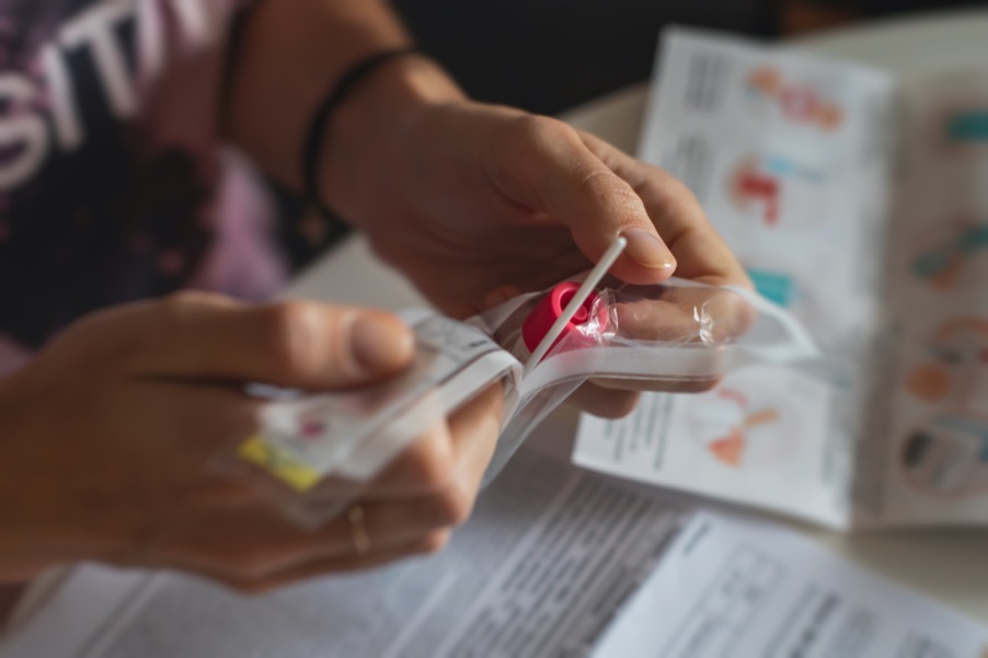 two hands are peeling open a plastic wrapping that holds a small plastic test tube with a round red cap, and a long white swab stick, a blurry instruction guide is in the background on a table.