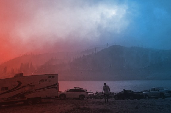 an eery landscape shot of a lake and nearby mountains except the sky is blotted around with thick orange smoke clouds