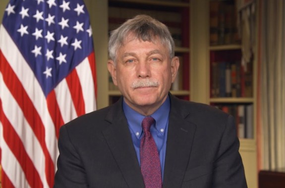 an older white man staring at the camera, with an office and an american flag behind him