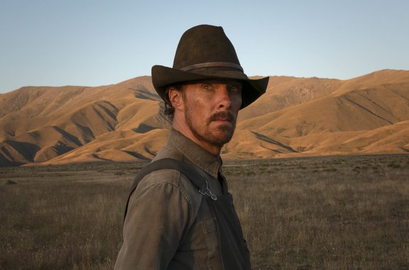 a white man in cowboy garb looks off camera over his shoulder with mountains in the background