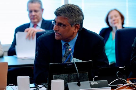 an older indian man in a suit tie in a meeting room sitting a desk, with an older white man and woman, also in suits, sitting behind him. a small microphone is in front of him as he looks to his right