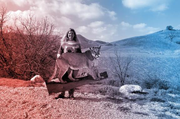 a middle aged white woman holding a cardboard cutout of a lion. a mostly flat desert landscape with a small mountain is behind her