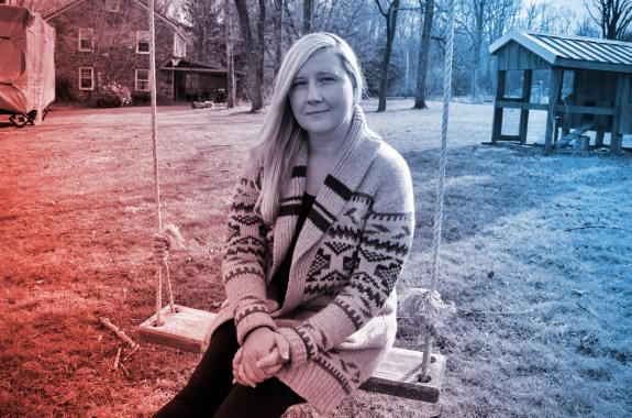 a white blonde-haired woman sitting on a swing outside looking pensive at the camera. behind her is grass, a shed, and a house