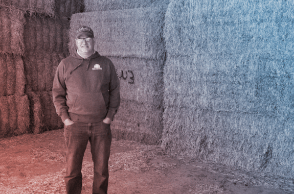 a man in a cap and sweater standing in front of bales of hay. the image is filtered blue and red