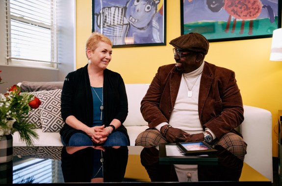 an older Black man and an older white woman talk to each other on a couch in a doctors waiting room