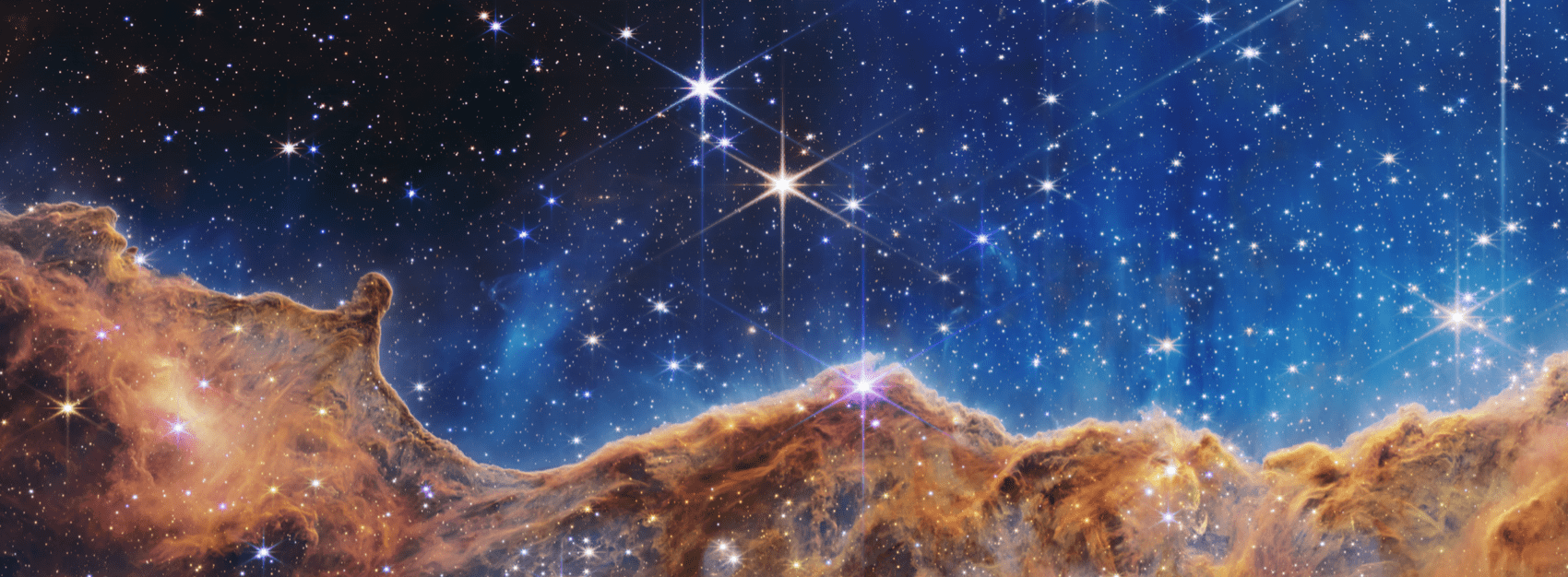 The image, taken by the JWST, is divided horizontally by an undulating line between a cloudscape forming a nebula along the bottom portion and a comparatively clear upper portion. Speckled across both portions is a starfield, showing innumerable stars of many sizes. The smallest of these are small, distant, and faint points of light. The largest of these appear larger, closer, brighter, and more fully resolved with 8-point diffraction spikes. The upper portion of the image is blueish, and has wispy translucent cloud-like streaks rising from the nebula below. The orangish cloudy formation in the bottom half varies in density and ranges from translucent to opaque. The stars vary in color, the majority of which, have a blue or orange hue. The cloud-like structure of the nebula contains ridges, peaks, and valleys – an appearance very similar to a mountain range. Three long diffraction spikes from the top right edge of the image suggest the presence of a large star just out of view.