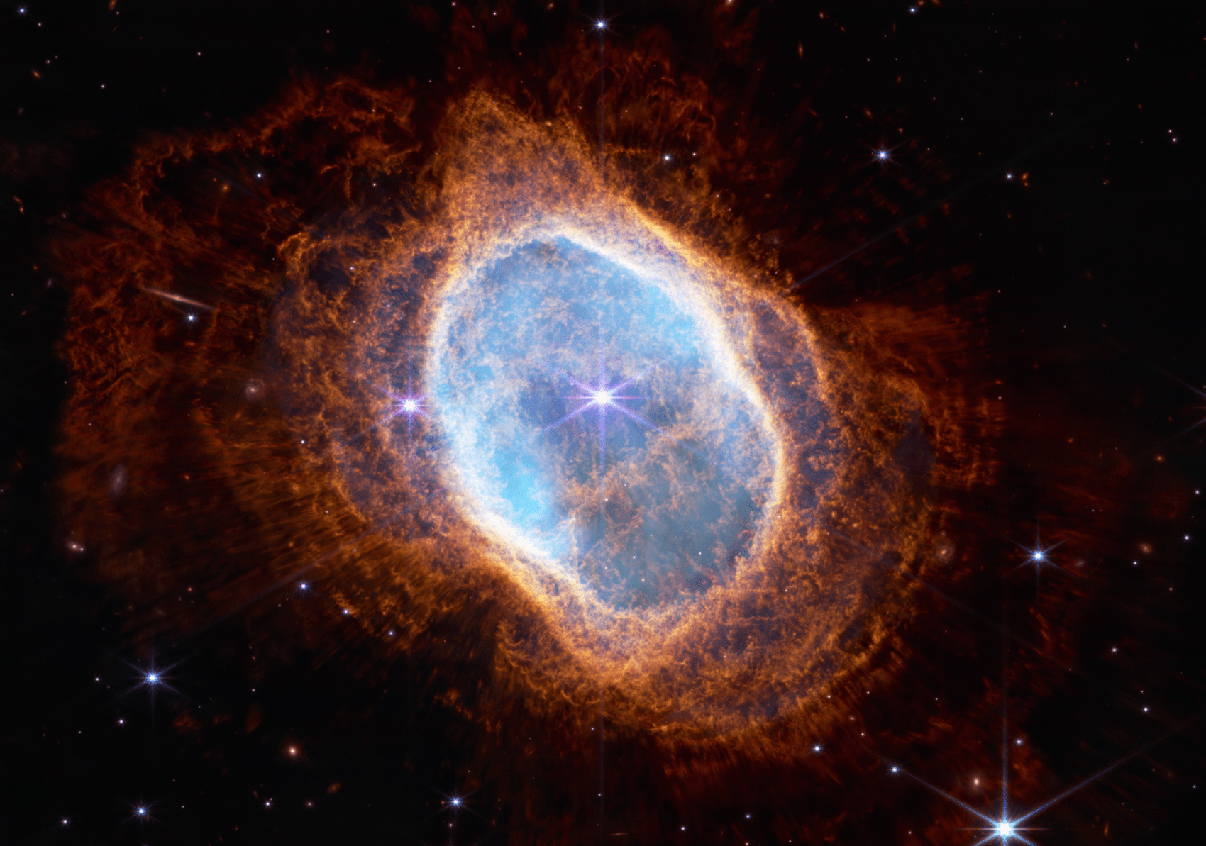 The image is split down the middle, showing two views of the Southern Ring Nebula. Both feature black backgrounds speckled with tiny bright stars and distant galaxies. Both show the planetary nebula as a misshapen oval that is slightly angled from top left to bottom right and takes up the majority of each image. At left, the near-infrared image shows a bright white star at the center with long diffraction spikes. Large, transparent teal and orange ovals, which are shells ejected by the unseen central star, surround it. At right, the mid-infrared image shows two stars at the center very close to one another. The one at left is red, the smaller one at right is light blue. The blue star has tiny triangles around it. A large transparent red oval surrounds the central stars. From that extend shells in a mix of colors, which are red to the left and right and teal to the top and bottom. Overall, the oval shape of the planetary nebula appears slightly smaller than the one seen at left.