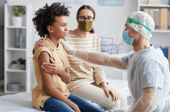 photo of a woman and middle school-aged child sitting side by side at a doctor's office, with a doctor reaching over to kindly touch the child's arm, while the child holds a piece of gauze to this other arm and smiles slightly