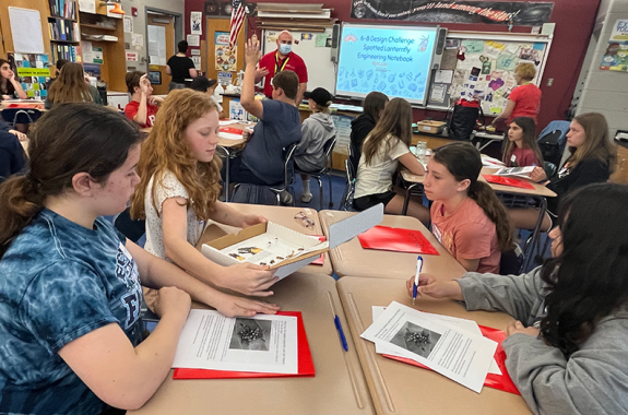 a photograph of students in a classroom working in groups of four at desks with boxes and worksheets featuring spotted lanternflies. the projection on the screen at the front of the class reads "6-8 Design Challenge Spotted Lanternfly Engineering Notebook"
