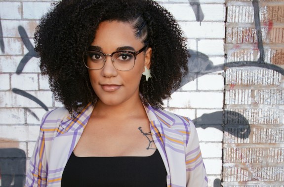 A woman with glasses looks into the camera with a thoughtful, gentle smile. A tattoo of a constellation peeks out from the right side of her shirt, by her collarbone.