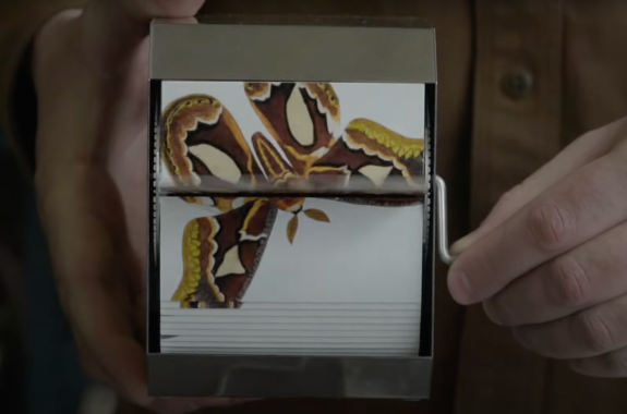a hand turns a crank on a small device the size of a napkin, flipping images of a brown moth in flight.