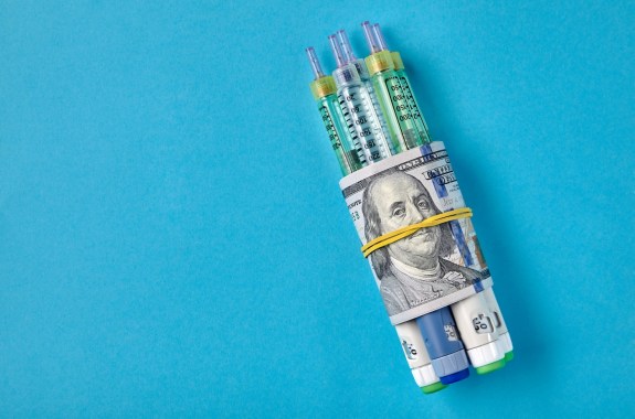 Insulin syringes wrapped in hundred dollar bills, blue background.