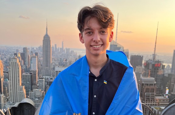 A smiling teen looking into the camera, wearing a uklranian flag around his shoulders with the NYC skyscape in the background.