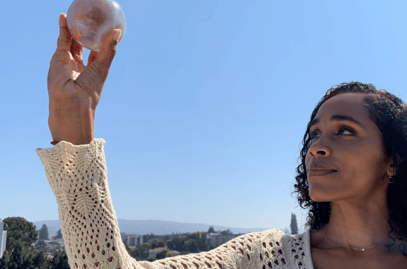 a black woman wearing a white long sleeve top holds up a 3d printed sphere the size of an apple that has the pattern of a marble