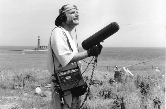 a black and white photo of an older white man wearing a backwards baseball cap with headphones and a large mic recorder over his shoulder, holds a large microphone with a wind cover on it as a seagull flies over his head