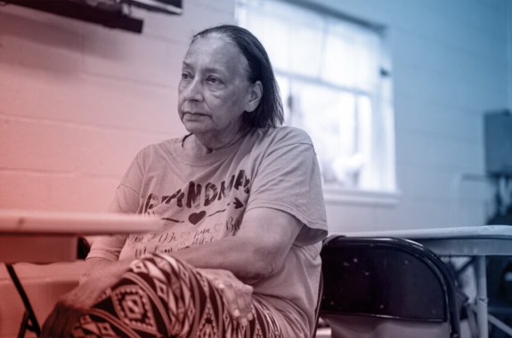 A middle-aged woman with a bright pink shirt sitting in a classroom, looking thoughtfully into the left of frame.