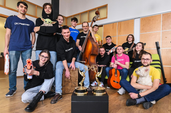 A team of 10 people smiling at the camera holding instuments, plush animals, and two Emmy awards.