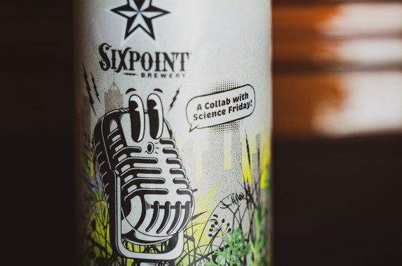 a closeup of a beer can. on the label is a old-school microphone with cartoon eyes looking to the left, it is also whistling. Behind it is a field of plants and a city skyline. the can says 'sixpoint brewery' and a speech bubble coming from the microphone says 'a collab with science friday!'