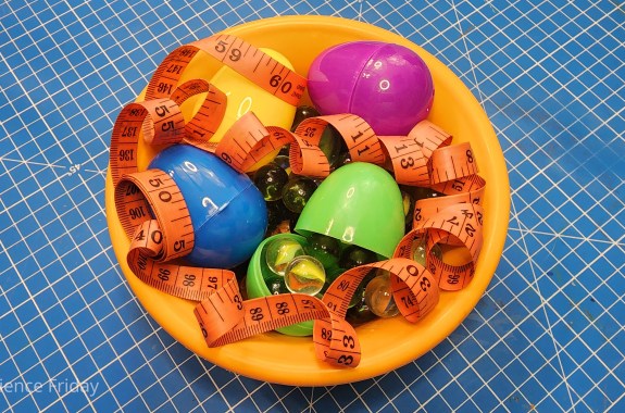 Materials for a science experiment. A bowl holds several plastic eggs, glass marbles and a tape measure.