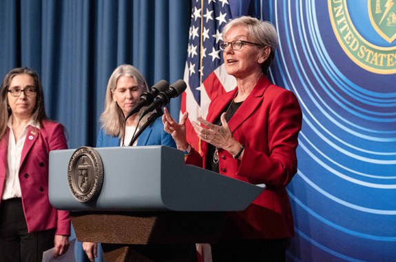 a press conference. an older white lady stands behind a podium speaking. to her right are two other white women looking on. an american flag and the department of energy logo are in the background