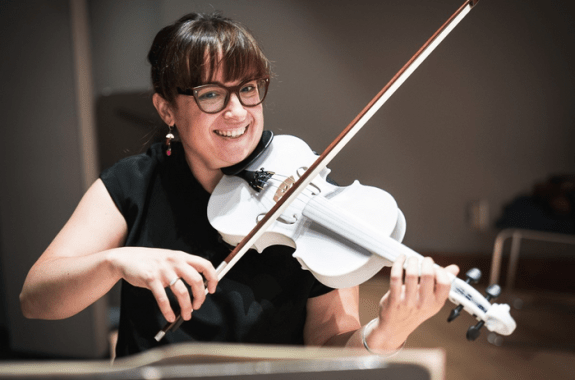 A woman smiling at the camera as she plays a bright white violin.