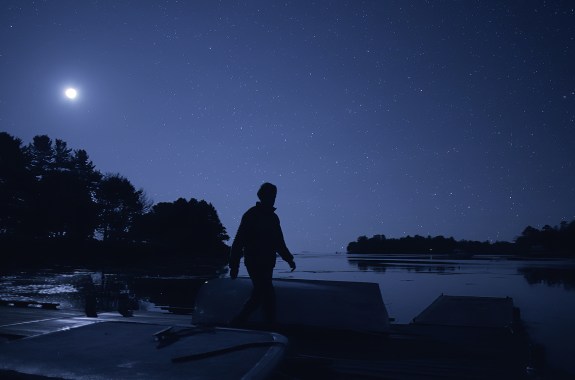 The silhouette of a man as he walks across a marshy landscape after the sun has set. Stars start to peek into view in the sky behind him.