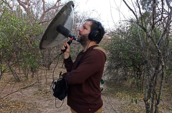 A man, Juan Pablo Culasso, holds a large metal dish towards the tops of trees, listening carefully to bird sounds.