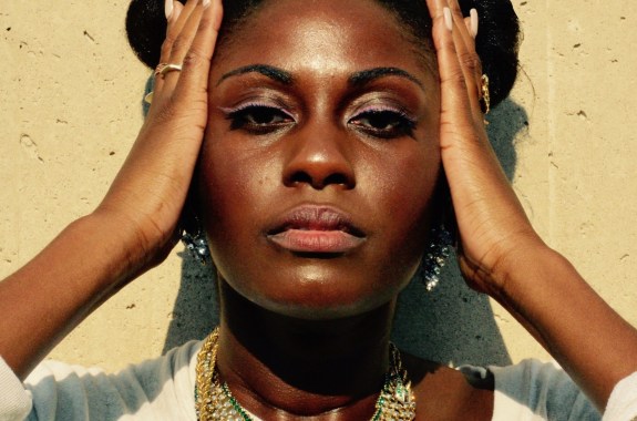 a Black woman against a wall holding her hands to the sides of her face looking directly at the camera