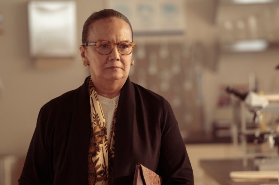 A woman in a small laboratory looks grimly toward the middle distance.