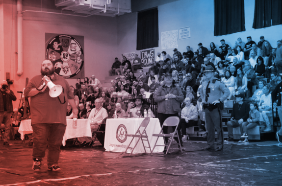 a man holding a megaphone walks in front of a crowd on bleachers and a cop seated in an auditorium