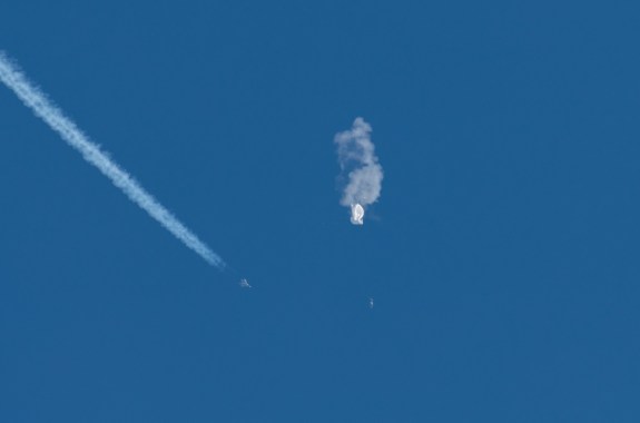 a zoomed in image of a deflated white balloon in the sky, and a fighter jet zooms by off to its left