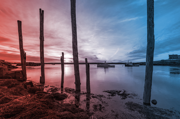 sunset at lowtide on the shore of a small coastal town in maine. a couple small ships sit on the water off in the distance. in the foreground are tall beams of wood, worn and misshapen after years of being stuck in the water