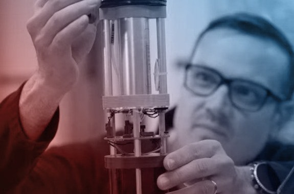 a red and blue filter over the image of a man in glasses holding a tube device that measures pressure in the ocean